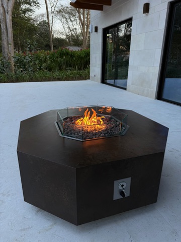 Proyecto custom FIREPITSCR: fire table hexagonal con acabado metálico oscuro, cristal antiviento y llamas al atardecer.