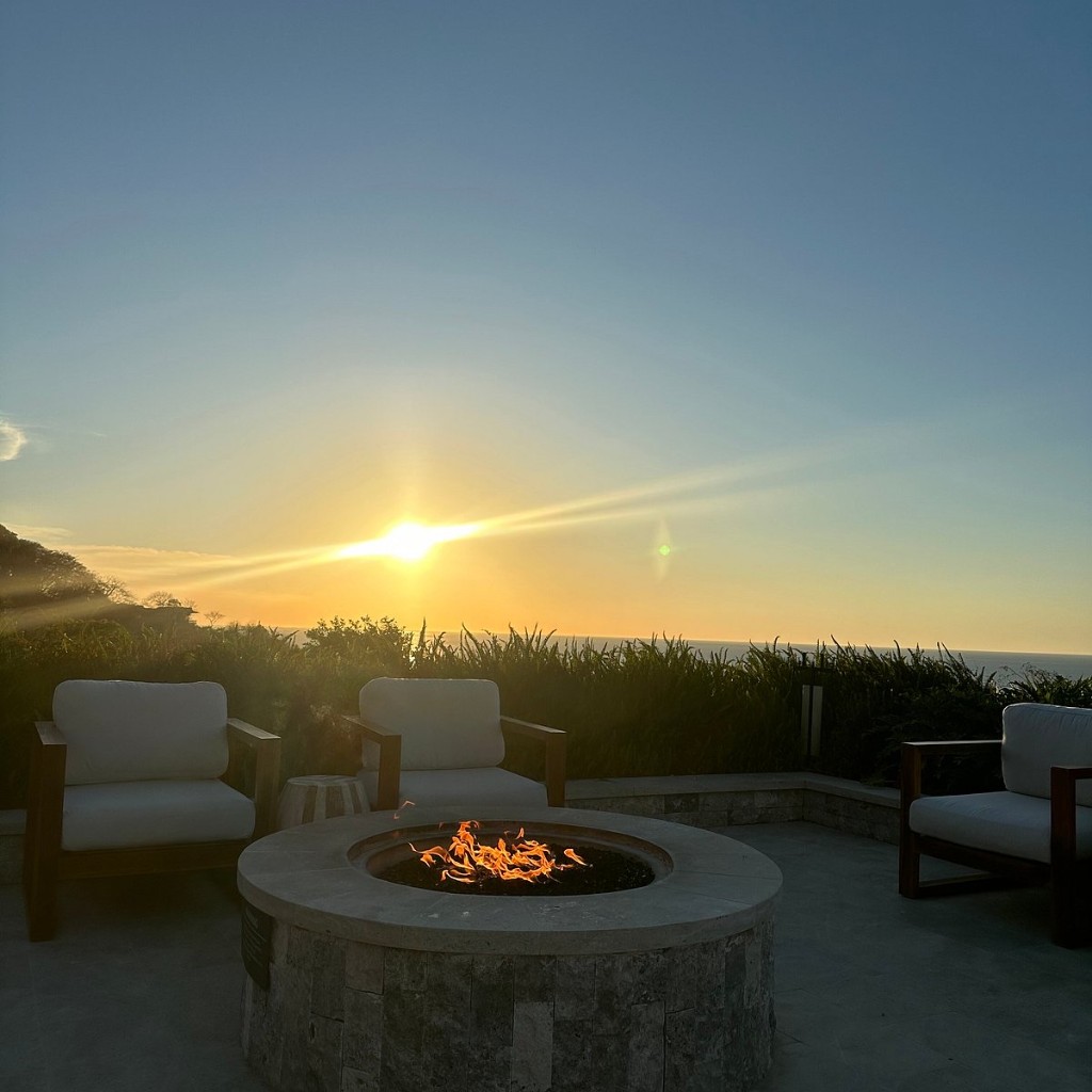 Firepit circular custom FIREPITSCR revestido en piedra clara, terraza frente al mar al atardecer.