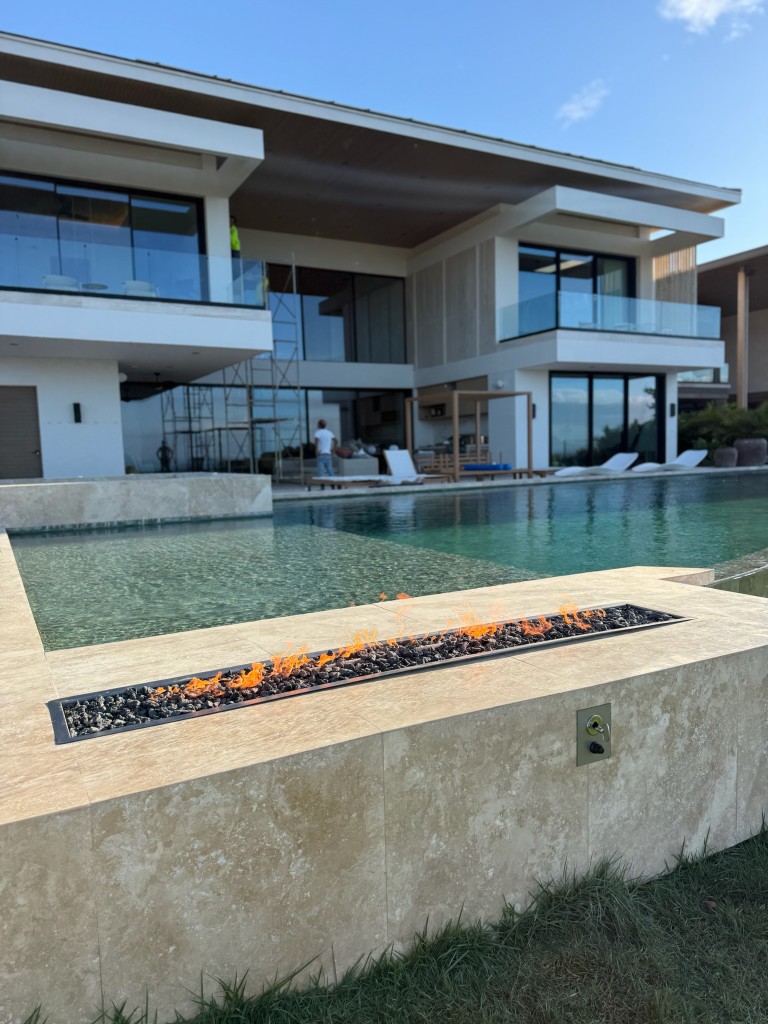 Hoguera lineal gas integrada en muro de travertino, proyecto custom FIREPITSCR junto a alberca y residencia moderna.