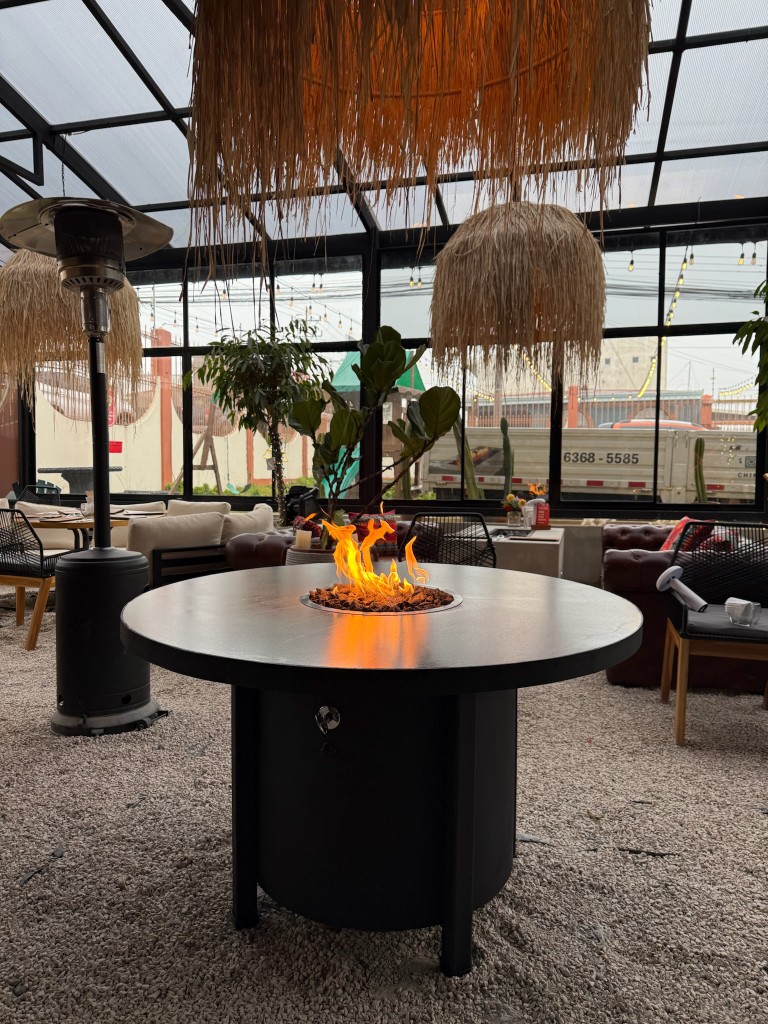 Mesa–fuego circular custom FIREPITSCR en metal negro mate, espacio lounge techado tipo invernadero.