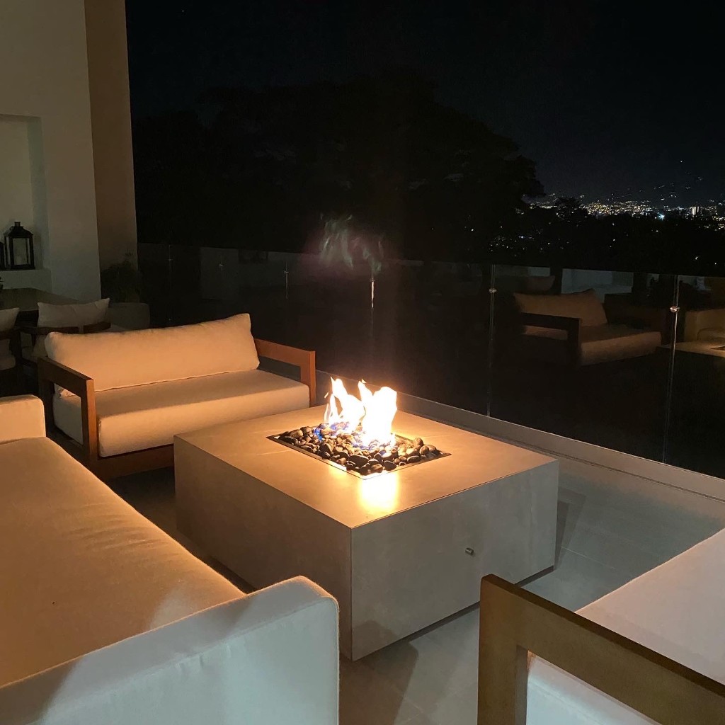 Fire table cuadrado FIREPITSCR de noche en terraza premium con sofás blancos y vista a luces de ciudad.