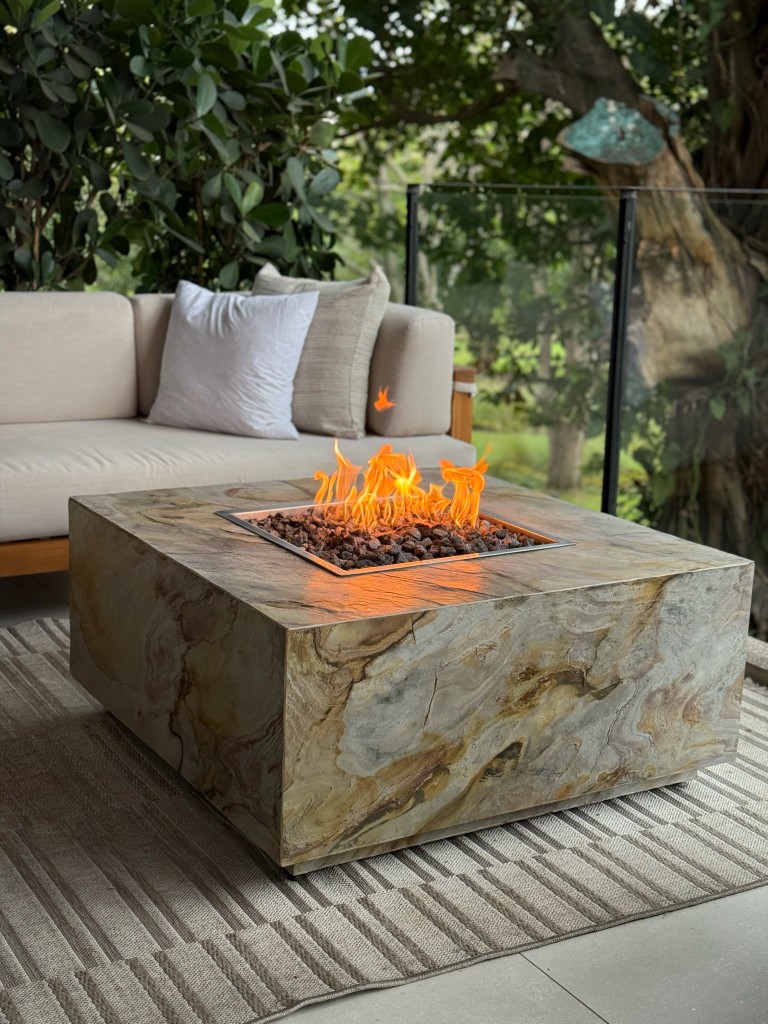 Fire table cuadrado FIREPITSCR con acabado tipo piedra vetada beige, llamas sobre lava en terraza con jardín.
