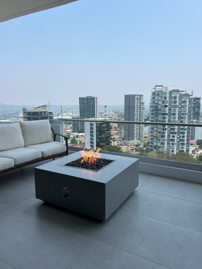 Fire table cuadrado FIREPITSCR en hormigón gris oscuro, balcón moderno con vista panorámica urbana.